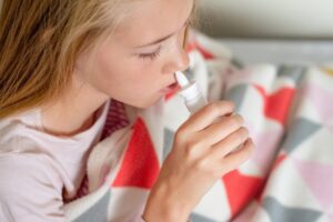 Sick teenage girl sitting on the bed covered with blanket using saline nasal spray